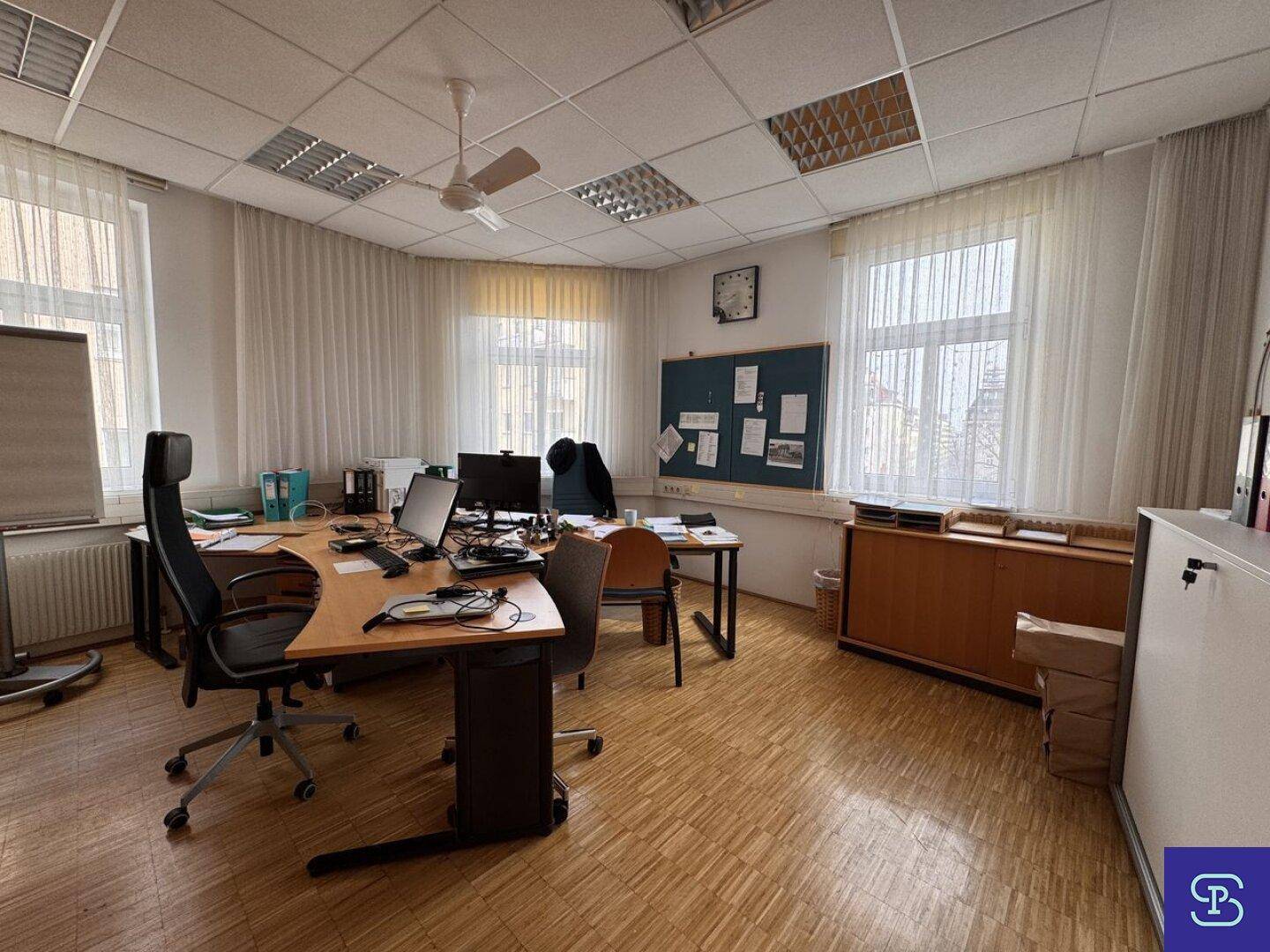 Büro mit Holzparkett, Deckenventilator und mehreren Arbeitsplätzen, mit großen Fenstern und Vorhängen.