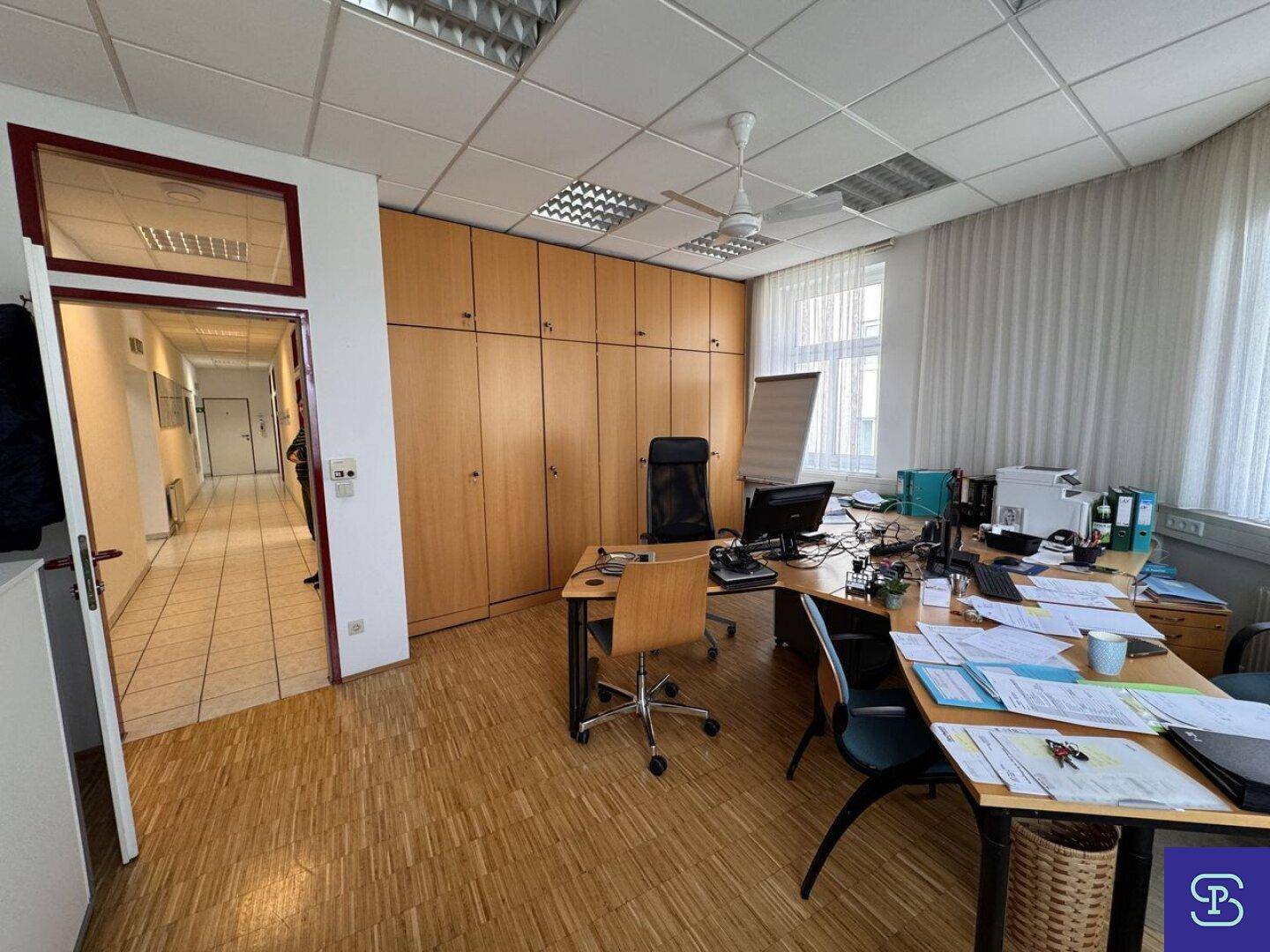 Büro mit Holzparkett, Deckenventilator und mehreren Arbeitsplätzen, mit Einbauschränken und Zugang zum Flur.