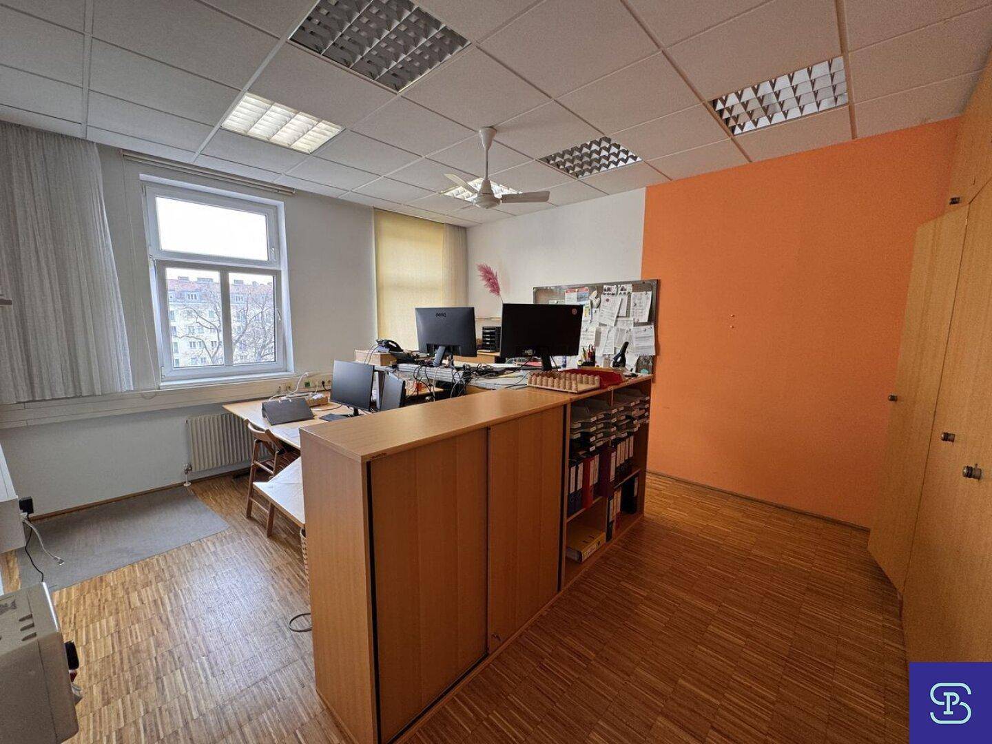 Büro mit Holzparkett, Deckenventilator und mehreren Arbeitsplätzen, mit orangefarbener Akzentwand.
