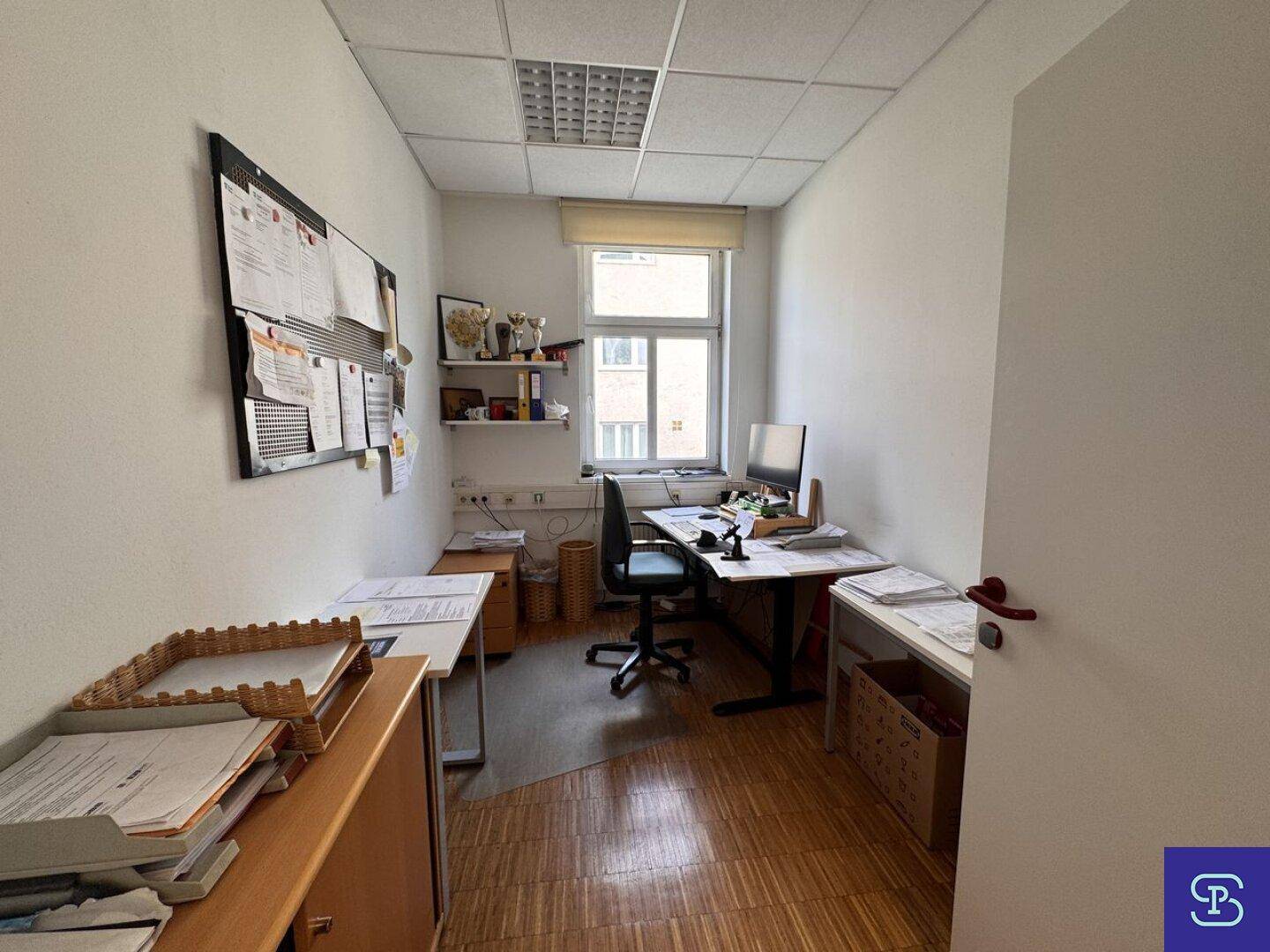 Kleines Büro mit Holzparkett und einem Arbeitsplatz, ausgestattet mit einem Fenster und Pinnwand.