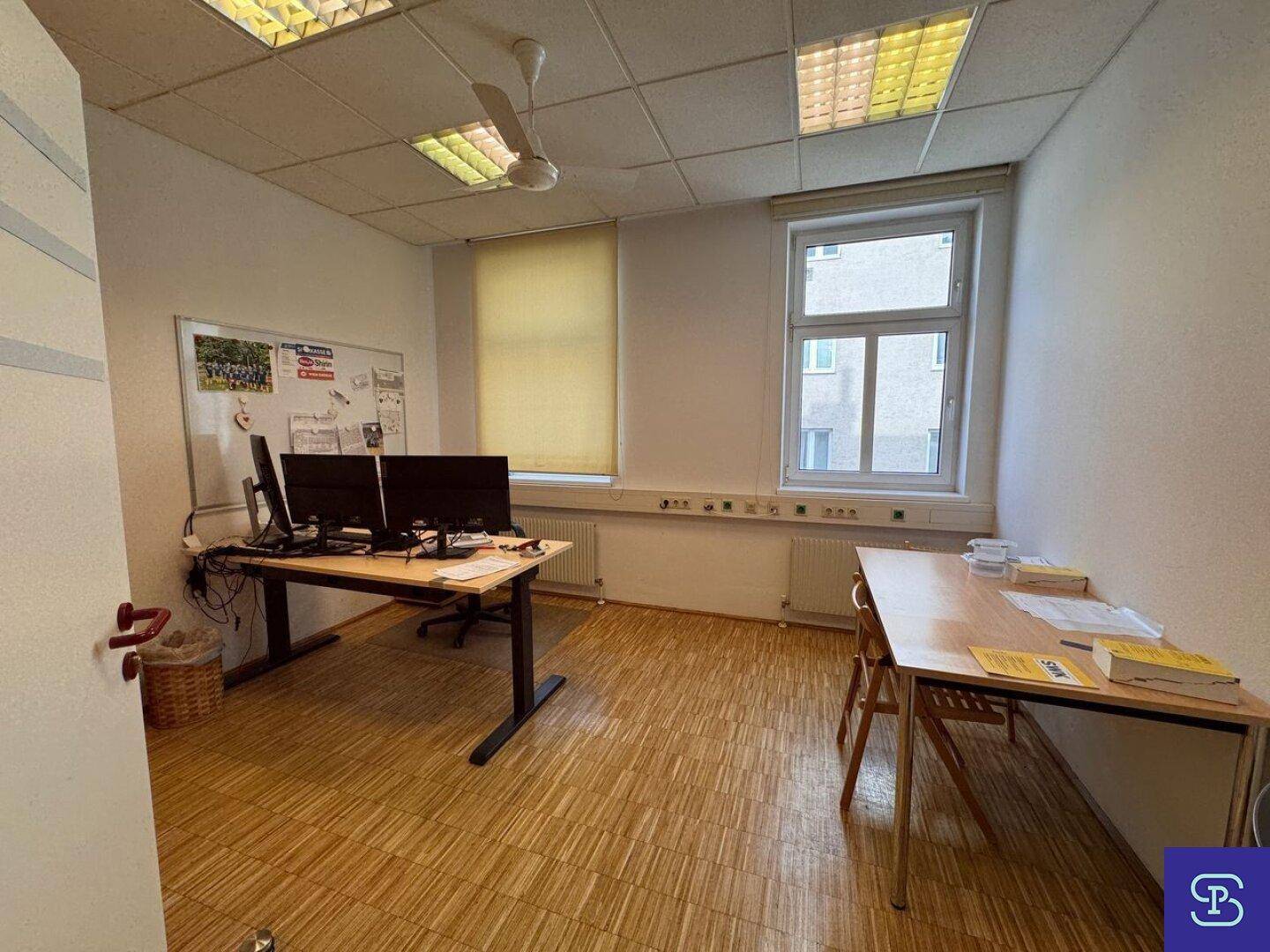 Büro mit Holzparkett, Deckenventilator und zwei Arbeitsplätzen, mit großen Fenstern und viel Tageslicht.
