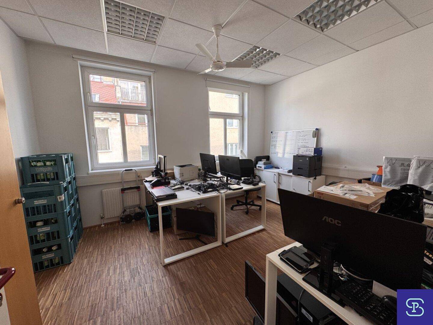 Büro mit Holzparkett, Deckenventilator und mehreren Arbeitsplätzen, mit großen Fenstern und Blick nach draußen.