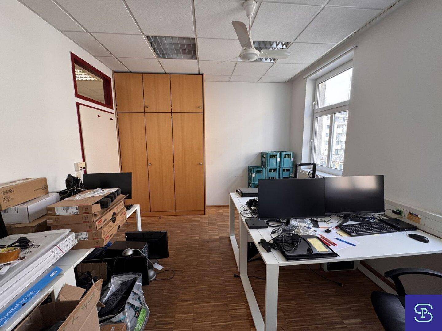 Büro mit Holzparkett, Deckenventilator und mehreren Arbeitsplätzen, mit großen Fenstern und viel Stauraum.