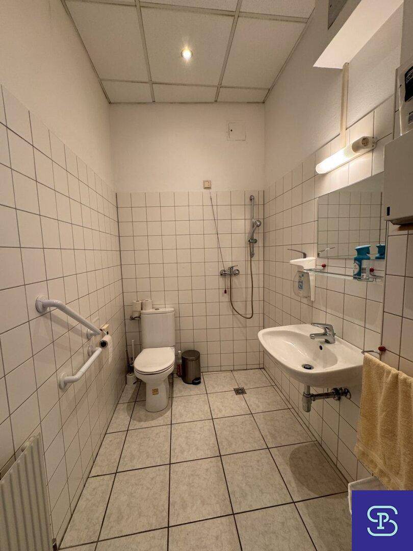 Badezimmer mit weißen Fliesen, Dusche und WC, ausgestattet mit Haltegriffen für Barrierefreiheit.