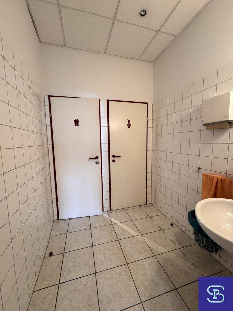 Badezimmer mit weißen Fliesen, Waschbecken und zwei Toilettenkabinen, sauber und funktional.