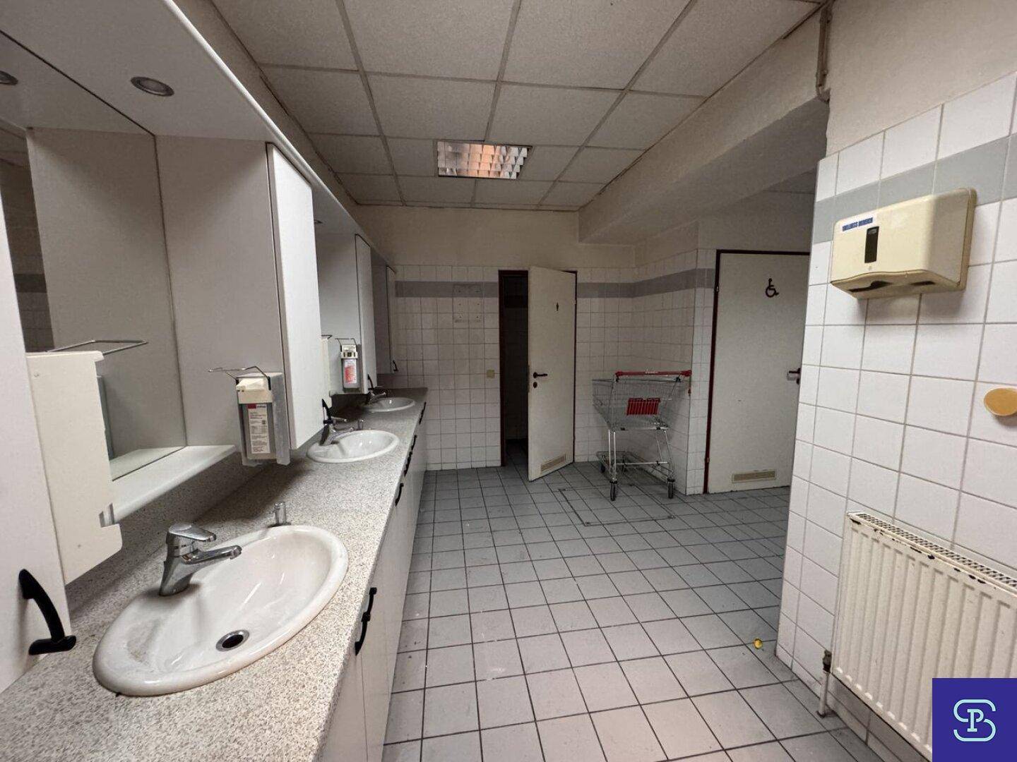 Großer Waschraum mit Fliesenboden, mehreren Waschbecken und Spiegeln, mit Zugang zu Toiletten.
