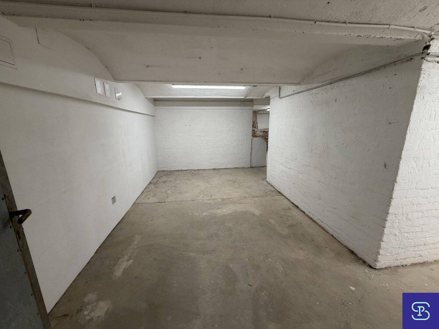 Langer Kellerraum mit weiß gestrichenen Ziegelwänden und Betonboden, beleuchtet durch Neonröhren.