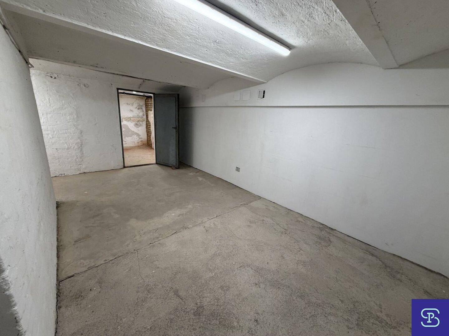 Kellerraum mit Betonboden und weiß gestrichenen Wänden, eine offene Tür führt zu einem weiteren Bereich.