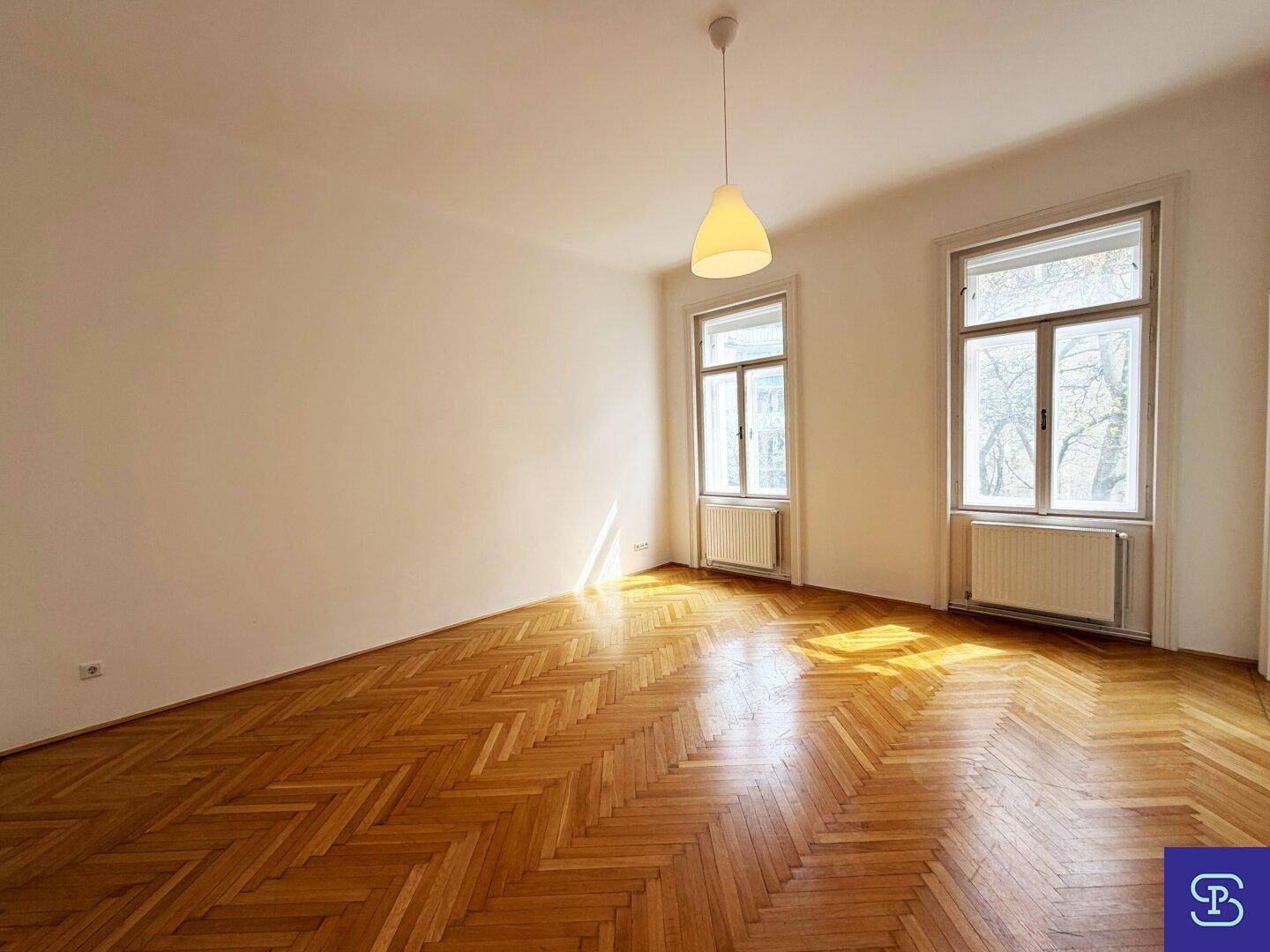 Helles Zimmer mit klassischem Fischgrätparkettboden, weißen Wänden und zwei großen Fenstern.