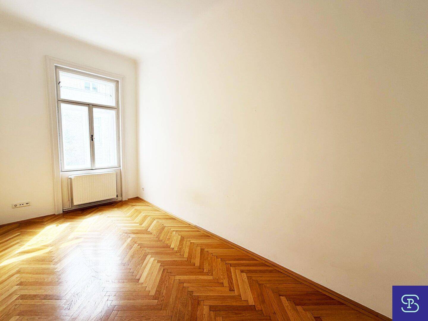 Geräumiger, leerer Raum mit Fischgrätparkettboden und einem großen Fenster, das viel Licht spendet.