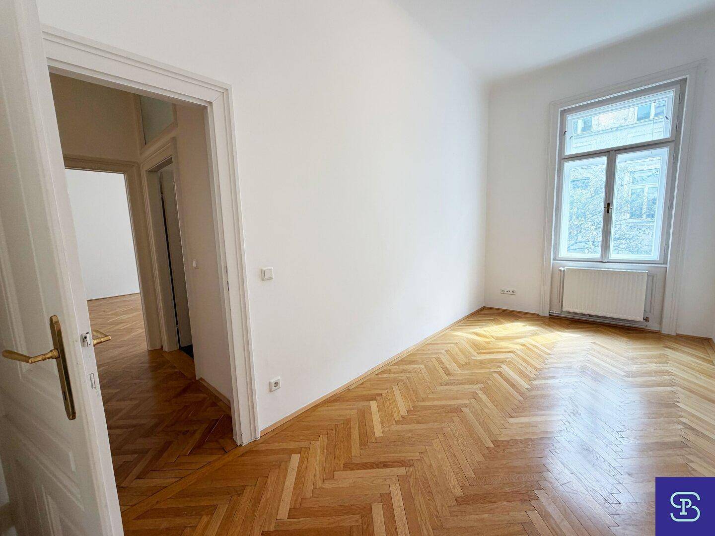 Geräumiges Zimmer mit Fischgrätparkett, weißen Wänden und einem großen Fenster mit Heizkörper.