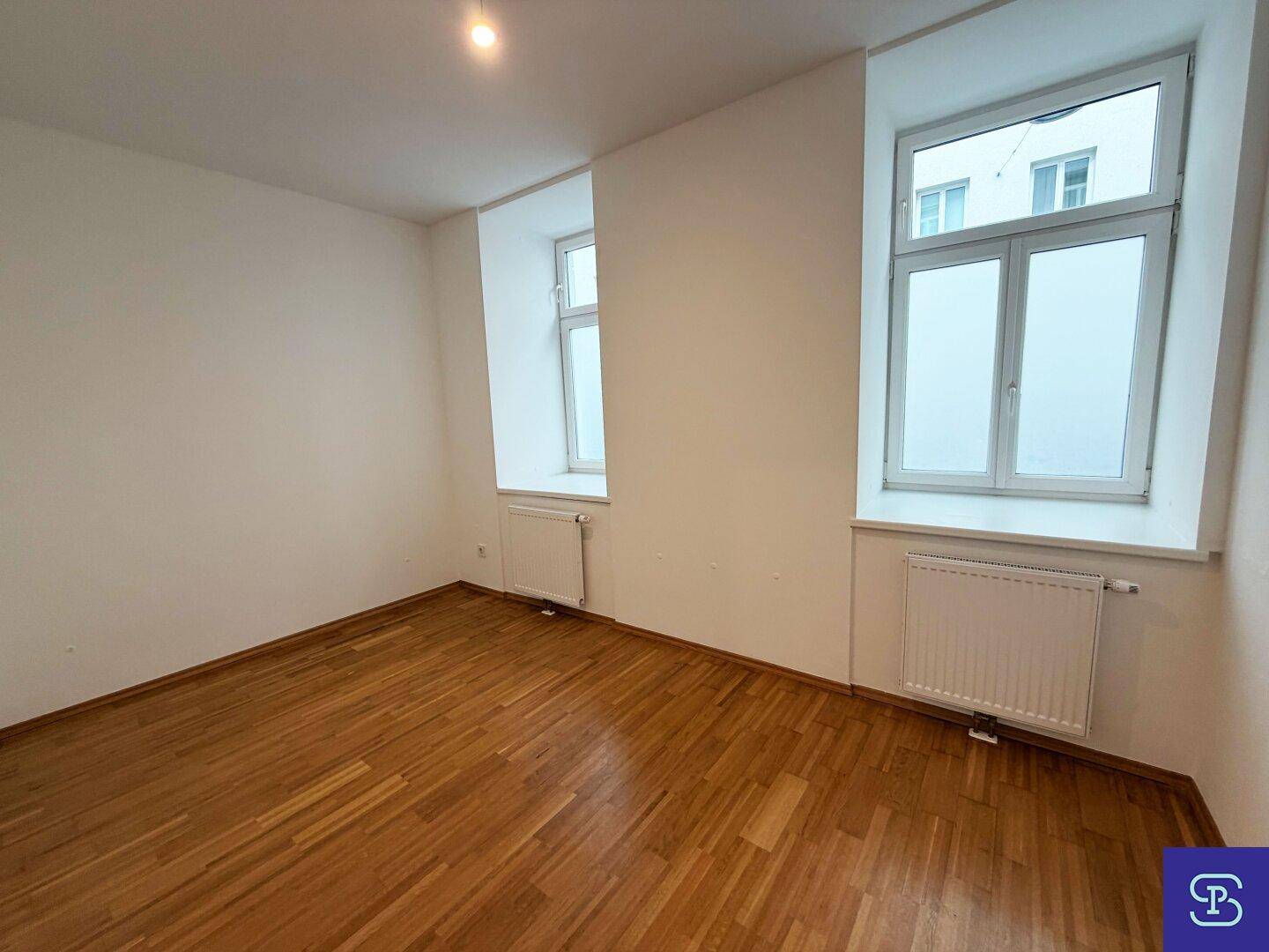 Helles Schlafzimmer mit zwei Fenstern, Parkettboden und Heizkörpern unter den Fensterbänken.