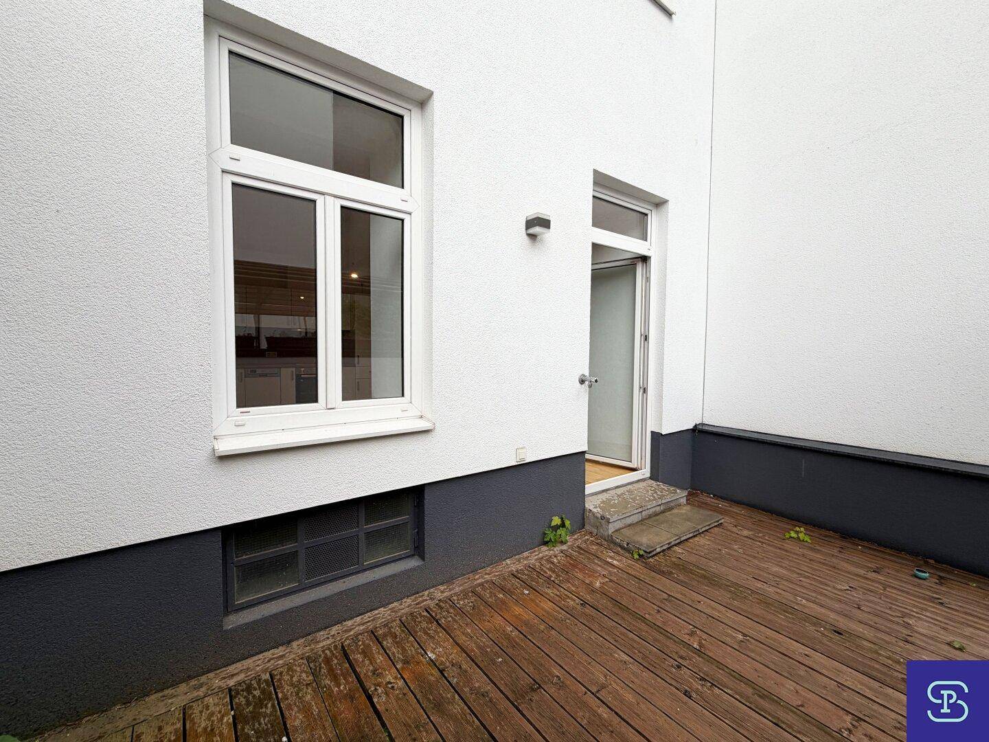 Kleiner Holzdeck-Patio mit Zugang vom Innenbereich und Blick auf die Gebäudefassade.