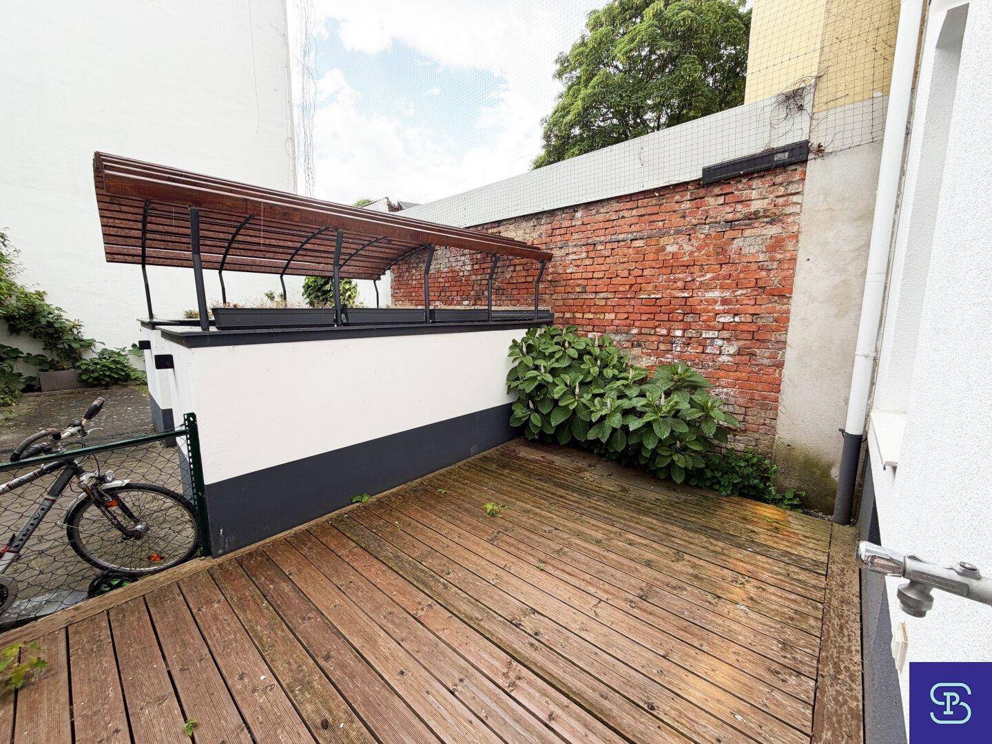 Kleiner Außenbereich mit Holzdeck, umgeben von einer Ziegelwand und etwas Grün, ideal zum Entspannen.