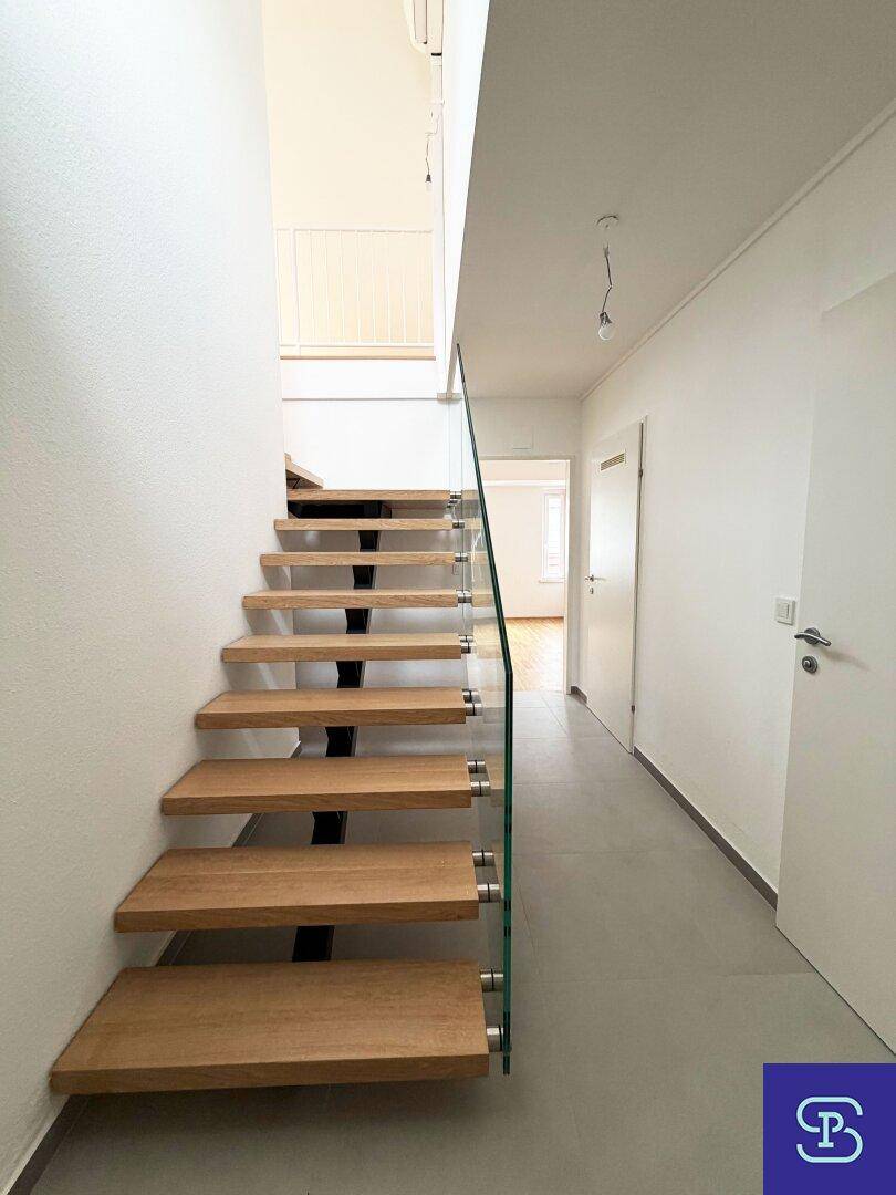 Elegante Holztreppe mit Glasgeländer in einem hellen Flur mit modernem Fliesenboden.