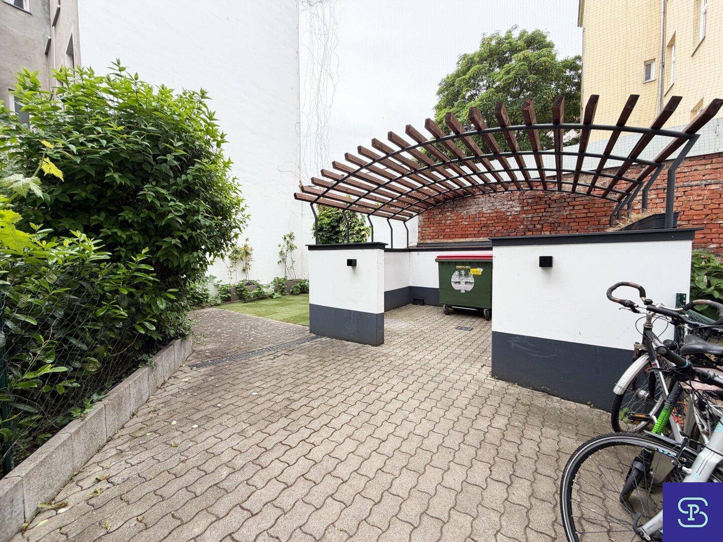 Gepflasterter Außenbereich mit Fahrradabstellplatz und überdachtem Mülltonnenbereich.