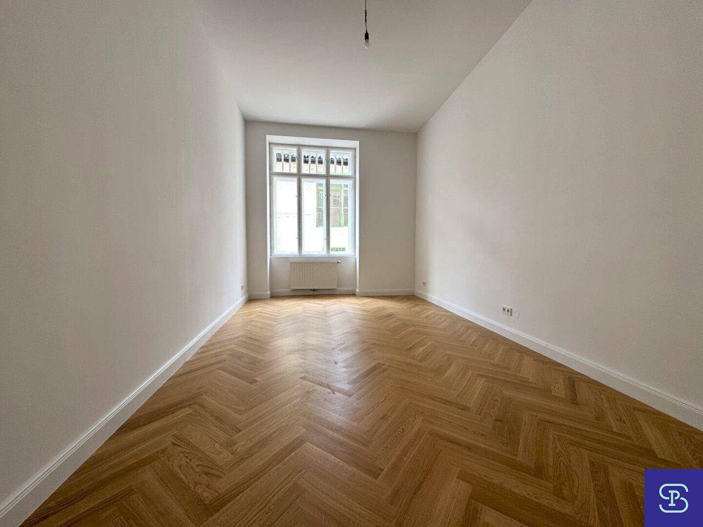 Helles Zimmer mit Fischgrätparkett und einem großen Fenster, das viel Licht spendet.