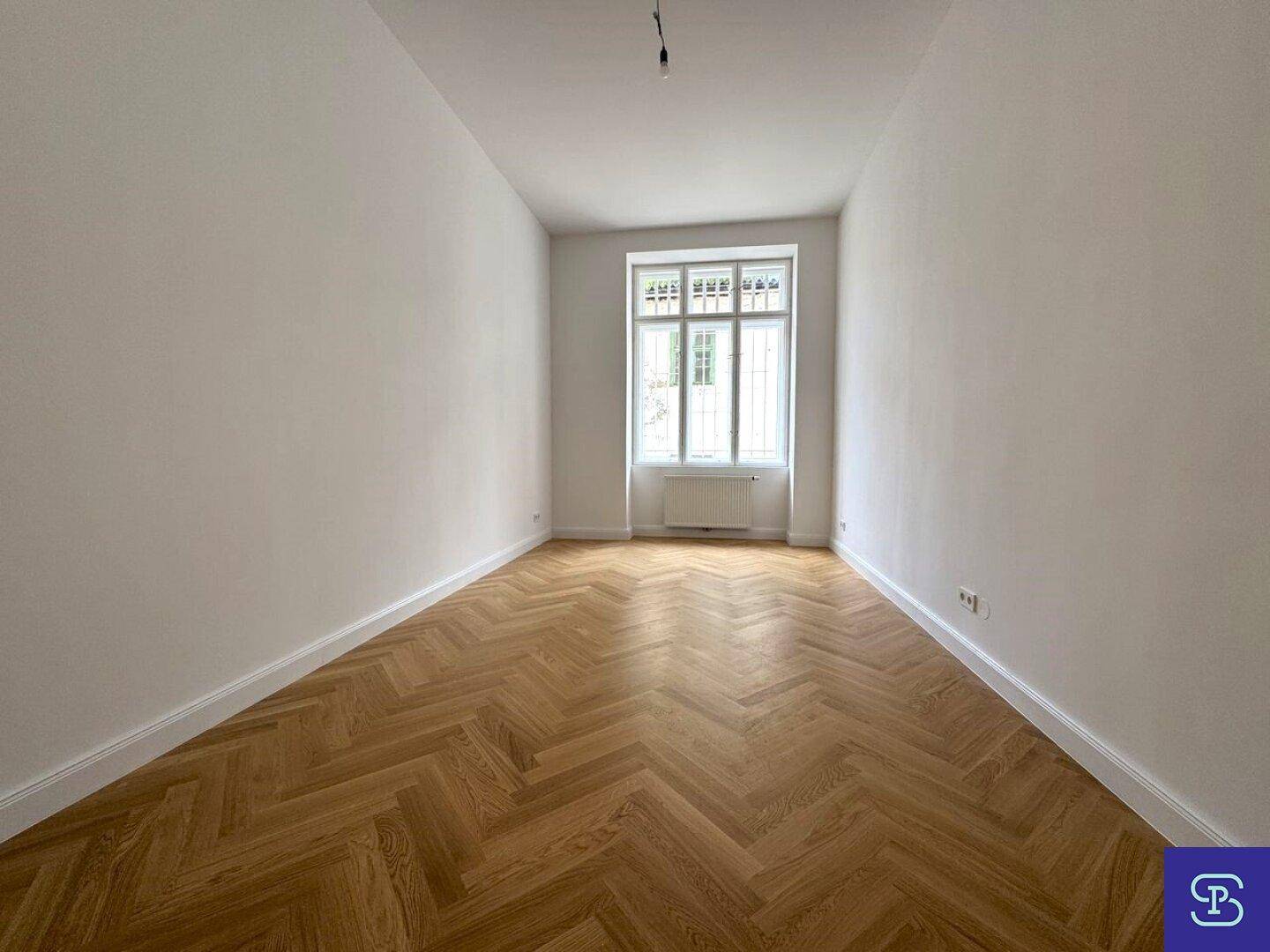 Langes, helles Zimmer mit Fischgrätparkett und einem großen Fenster.