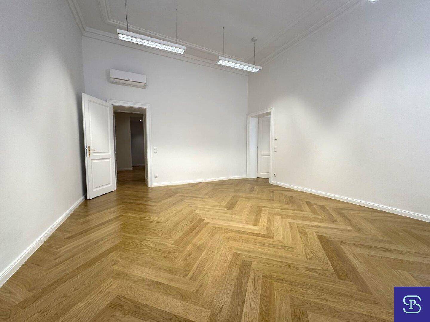 Heller Büroraum mit elegantem Fischgrätparkett, Klimaanlage und Zugang zu weiteren Zimmern durch weiße Türen.