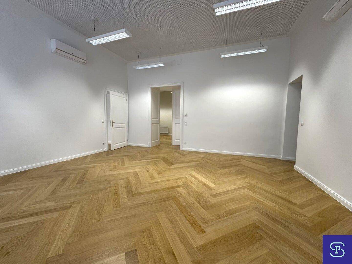 Leerer Büroraum mit Fischgrätparkett, hohen Decken und mehreren Türen, die zu anderen Bereichen führen.