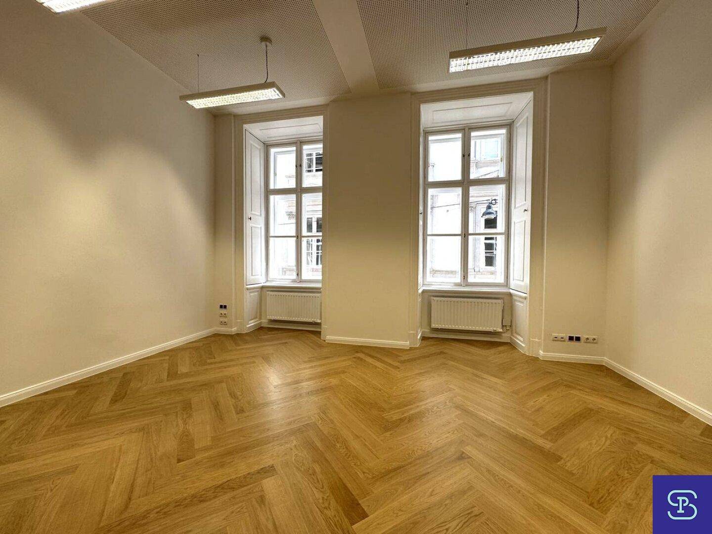 Großer Büroraum mit Fischgrätparkett, hohen Fenstern und Fensterläden, die viel Tageslicht hereinlassen.