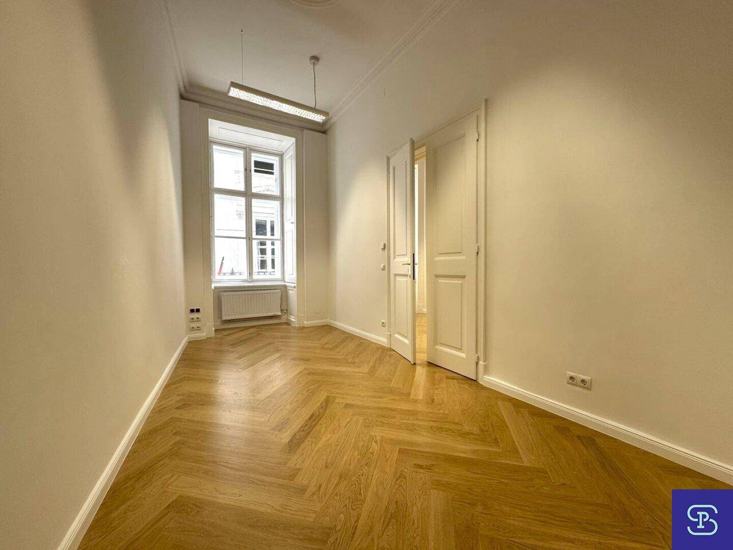 Kleinerer Büroraum mit Fischgrätparkett, hohen Fenstern und einem einzelnen Deckenlicht, gut beleuchtet.