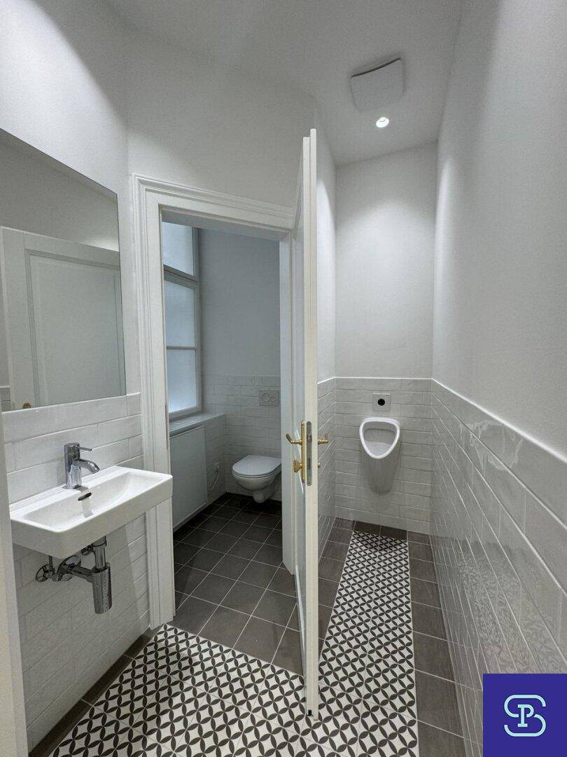 Helles Gäste-WC mit Urinal, Waschbecken und Fenster, das in einen weiteren Toilettenbereich führt.