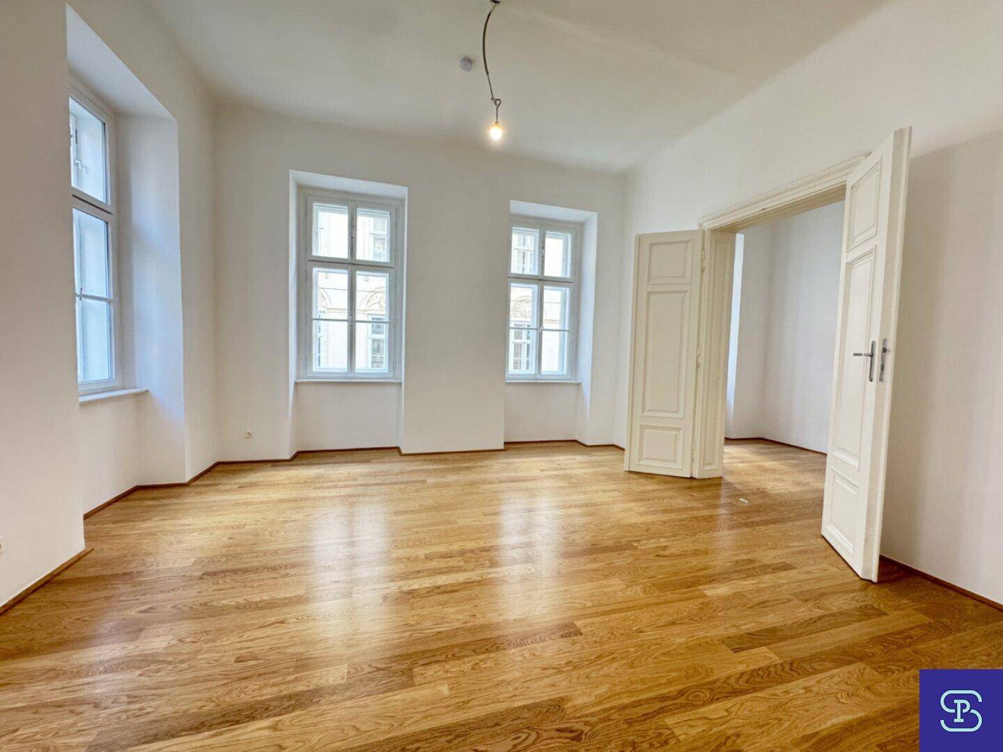 Geräumiges Wohnzimmer mit Holzboden und zwei hohen Fenstern, die viel Licht hereinlassen.