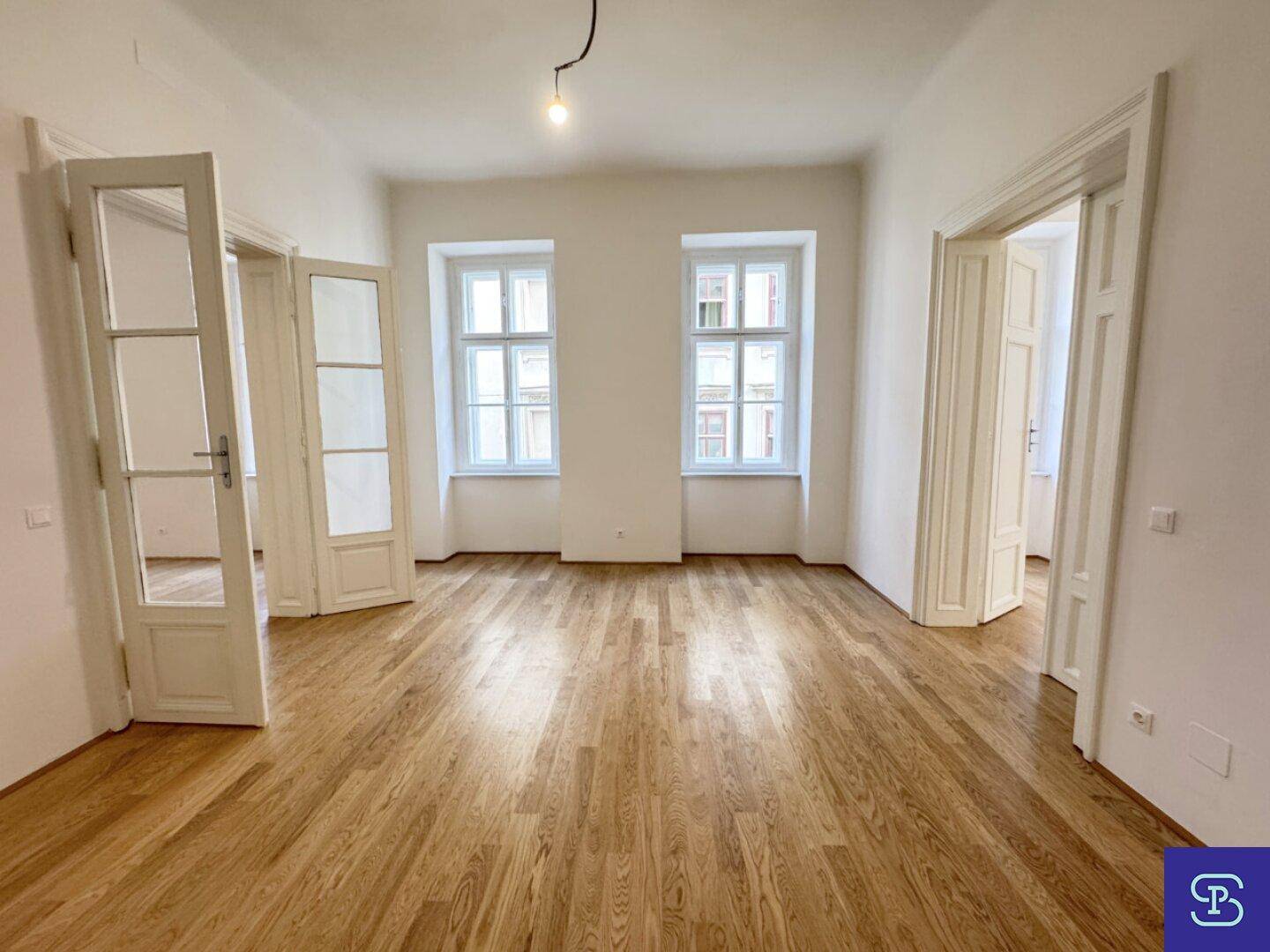 Geräumiges Wohnzimmer mit Holzboden und hohen Fenstern, offener Übergang zu anderen Bereichen.