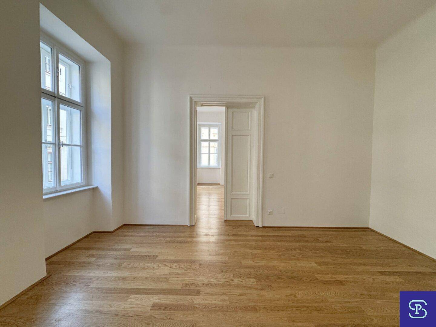 Großes, helles Wohnzimmer mit Holzboden und einem hohen Fenster, das viel Tageslicht bietet.