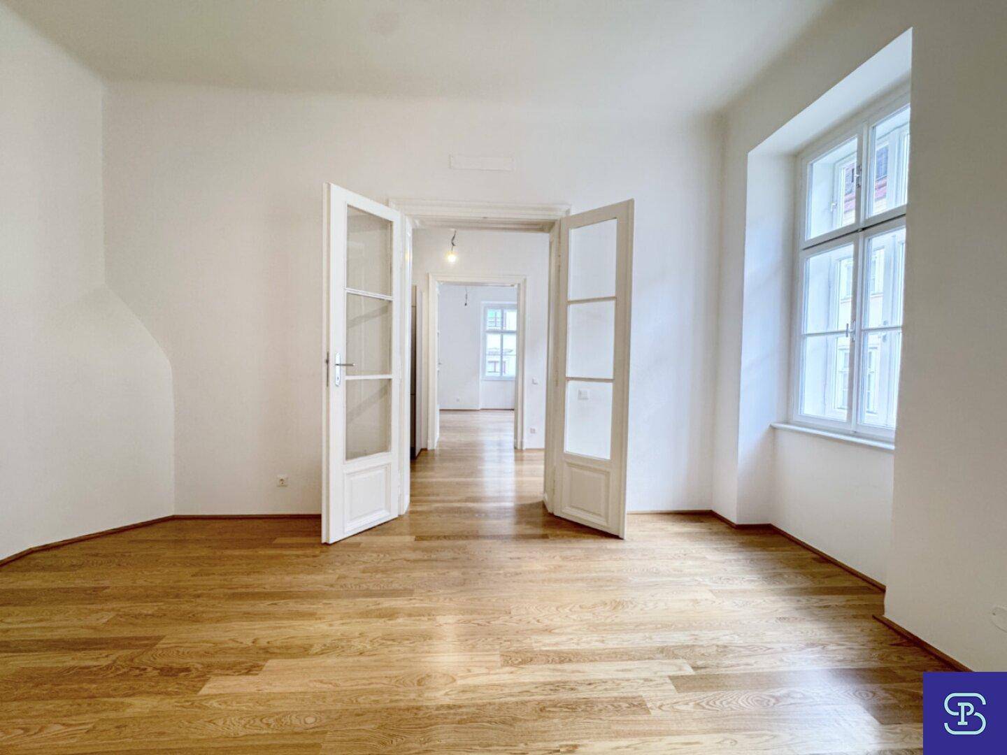 Heller Raum mit Holzboden und Doppeltüren, die den Blick in weitere Zimmer freigeben.