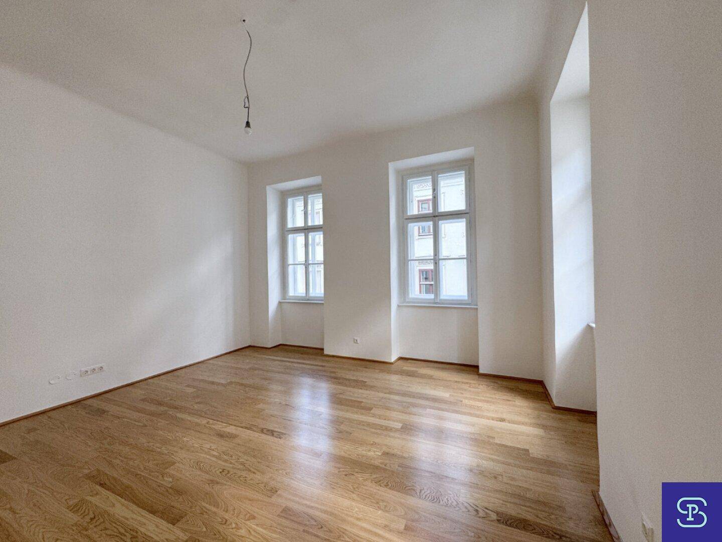 Helles Zimmer mit glänzendem Holzboden und zwei hohen Fenstern, die viel Licht spenden.