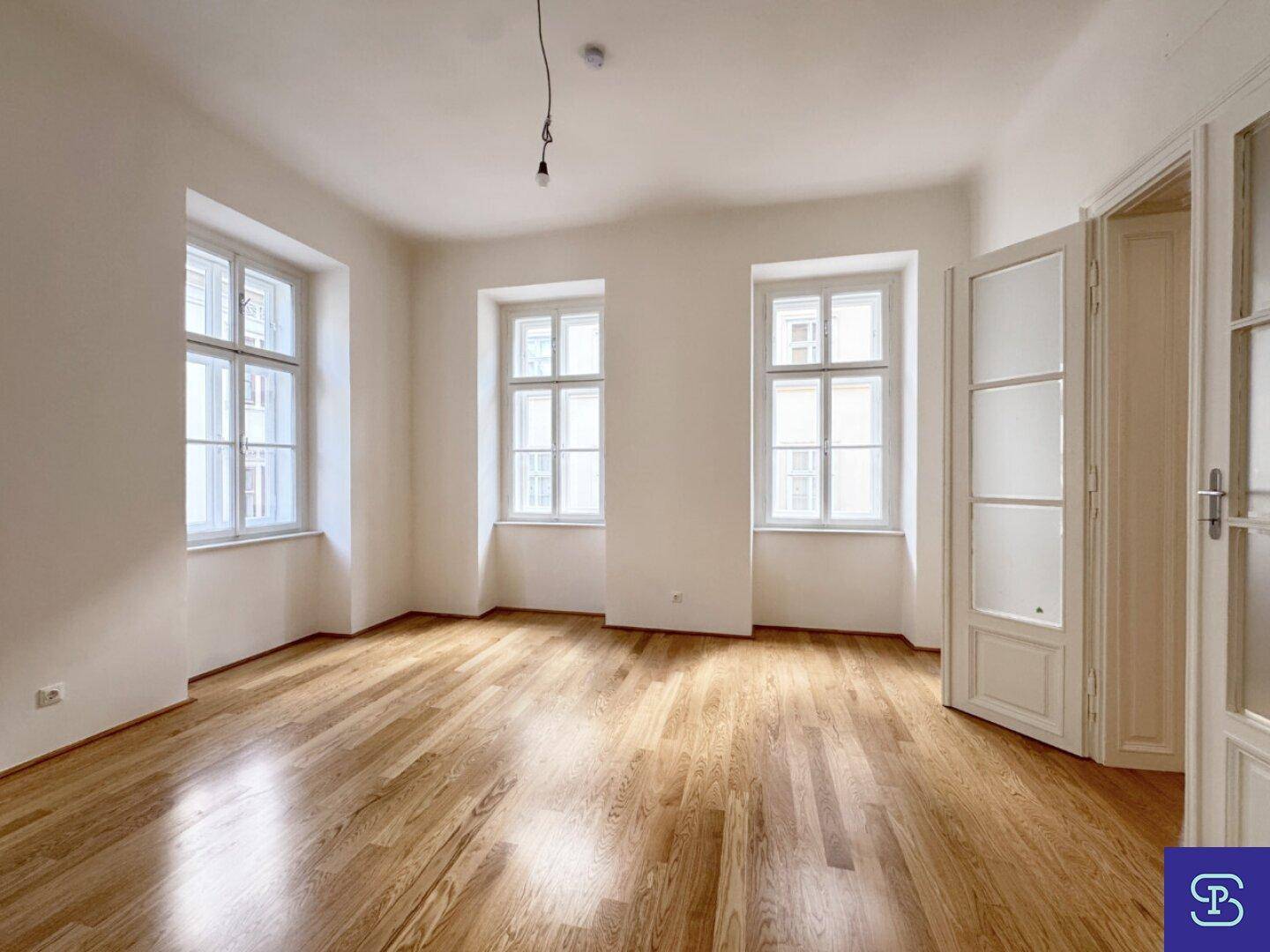 Lichtdurchflutetes Zimmer mit Holzboden, drei hohen Fenstern und eleganten Doppeltüren.
