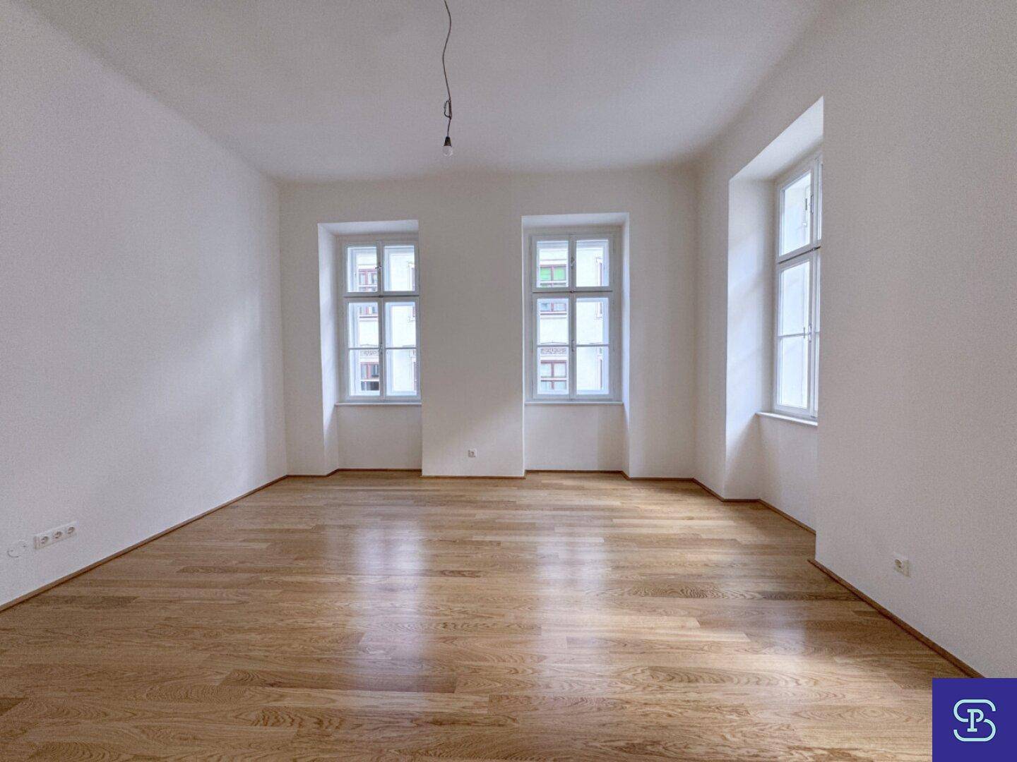 Geräumiges Zimmer mit Holzboden und drei hohen Fenstern, das eine helle Atmosphäre schafft.