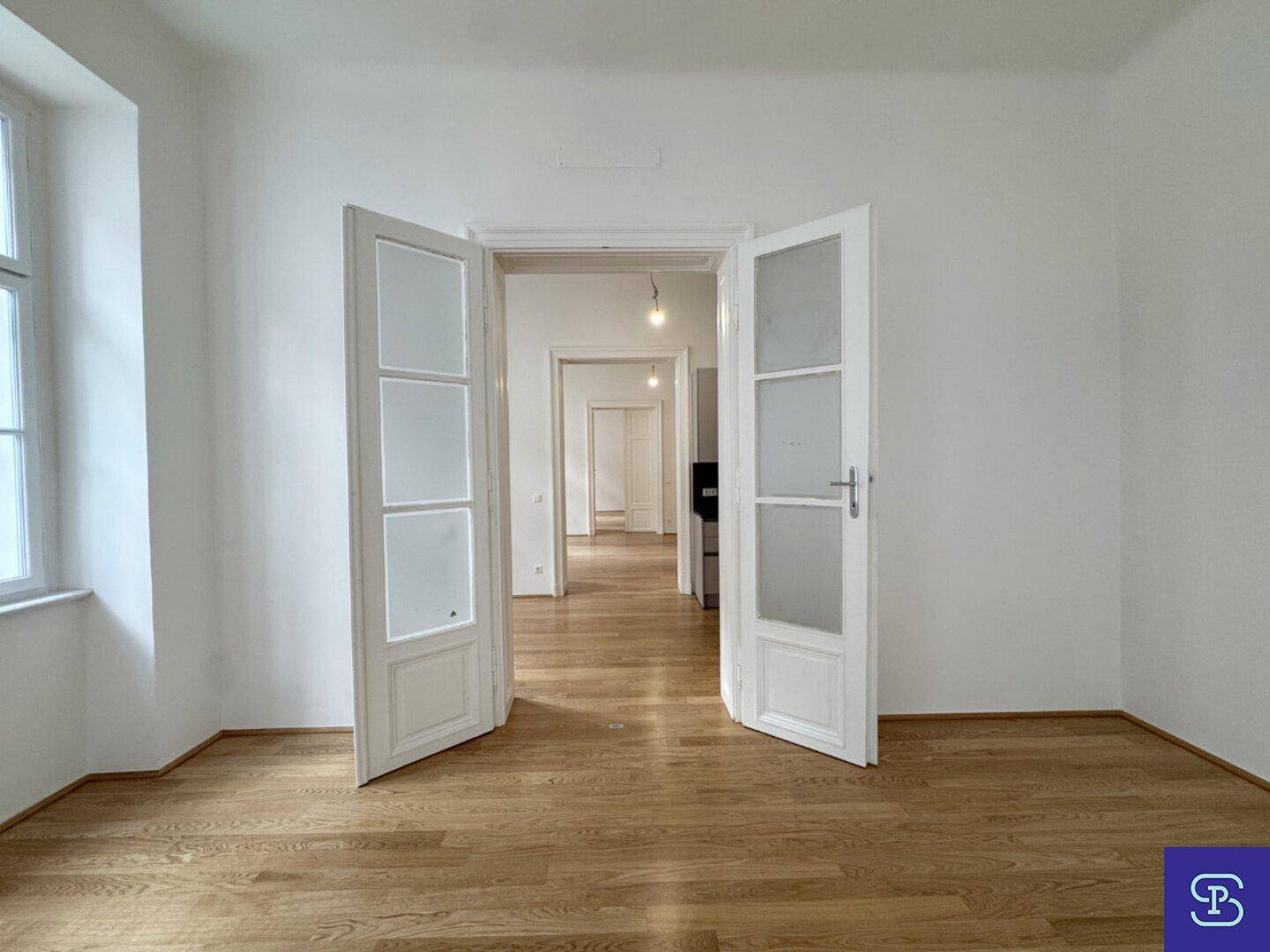 Heller Raum mit Holzboden und Doppeltüren, die den Blick in weitere Zimmer freigeben.