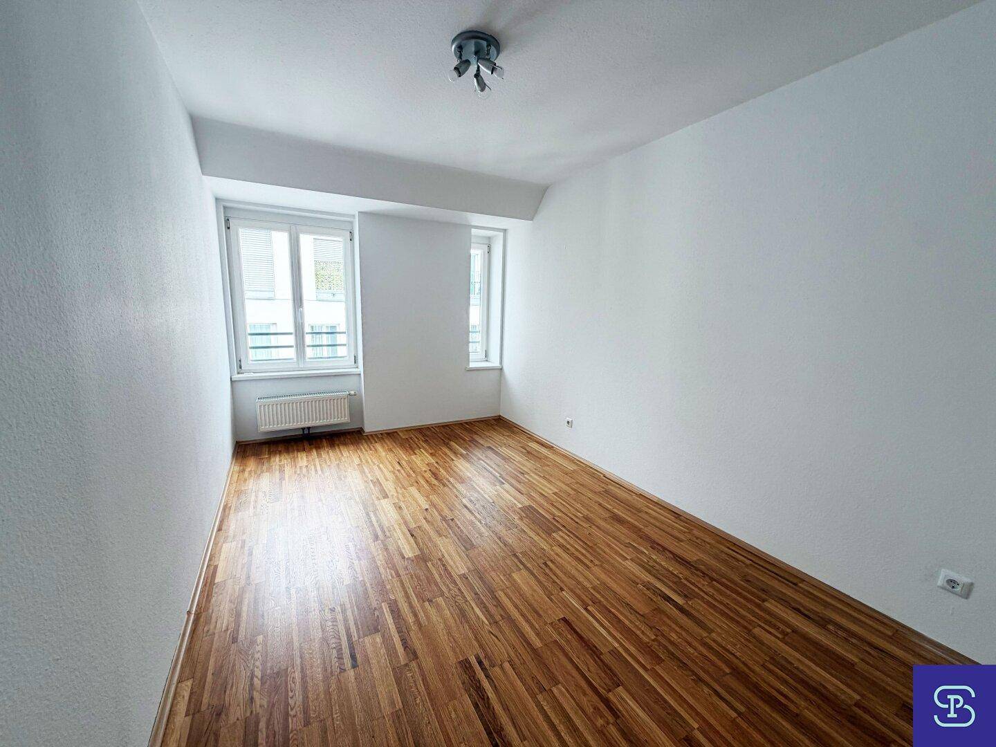 Leeres Zimmer mit Parkettboden und zwei Fenstern, bereit zur individuellen Gestaltung.
