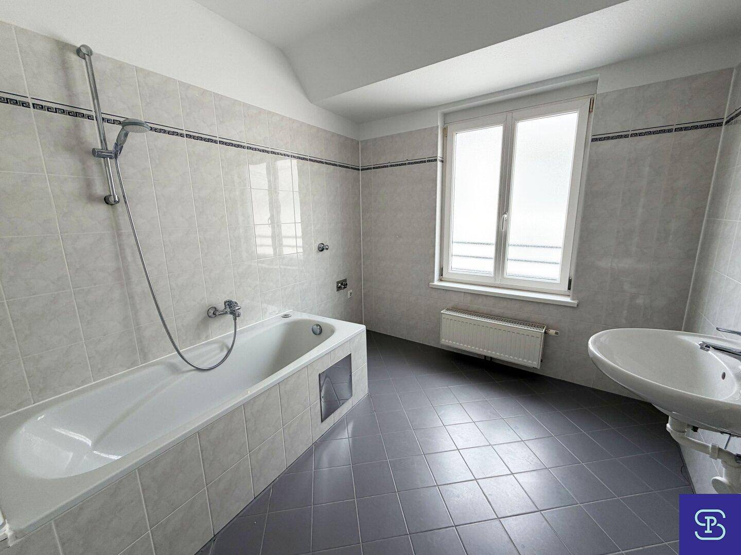 Gefliestes Badezimmer mit Badewanne, Waschbecken und Fenster, in hellem Design.