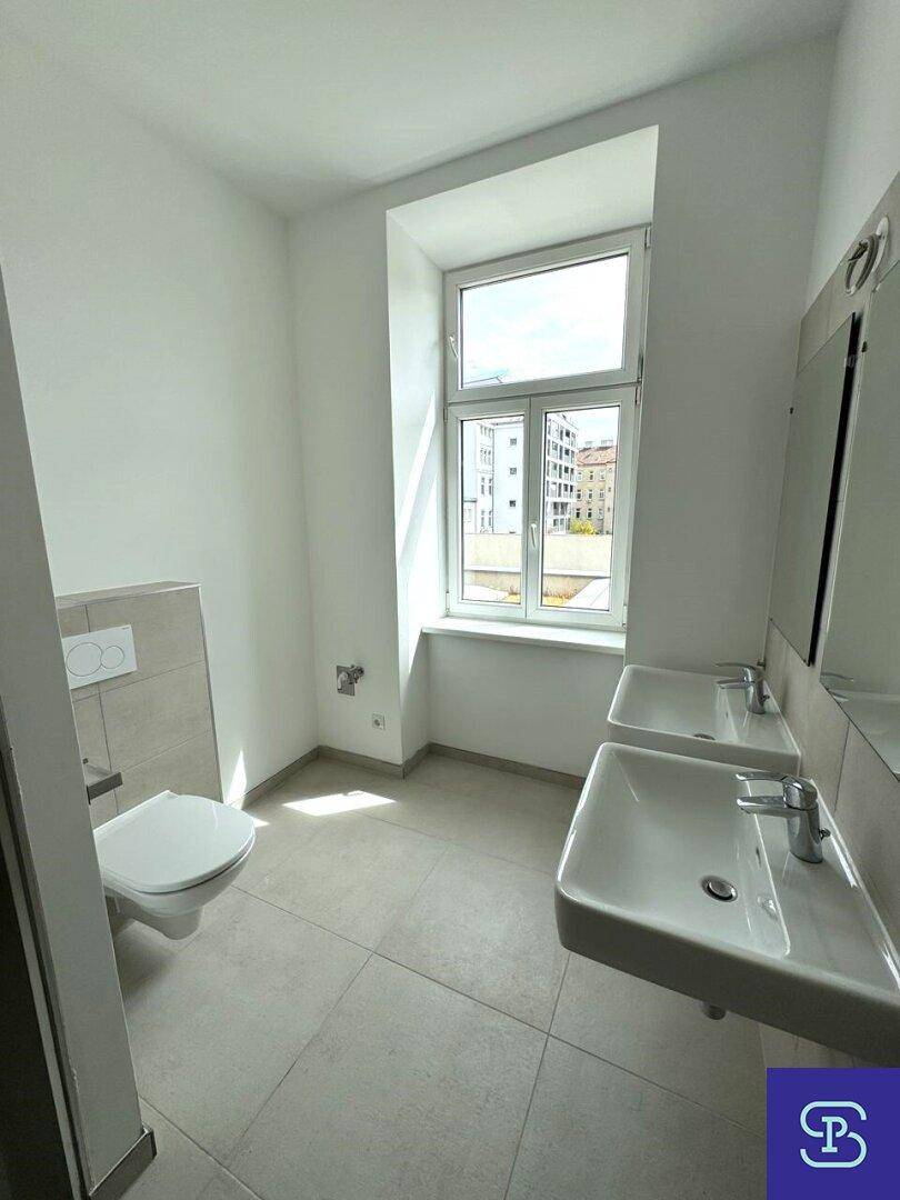 Helles Badezimmer mit Doppelwaschbecken, wandhängendem WC und großem Fenster.