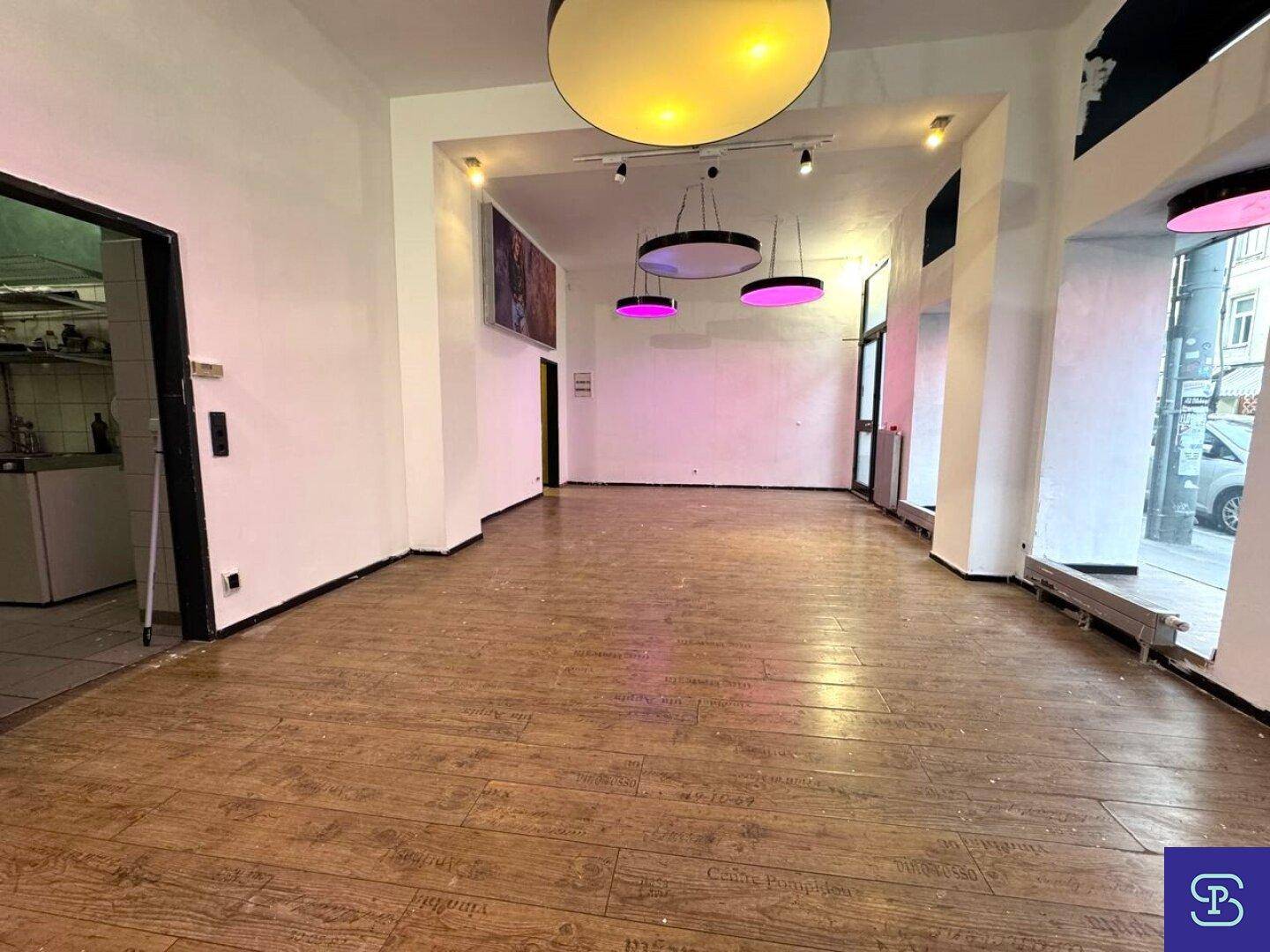 Blick in den leeren Ladenbereich mit Holzboden und Zugang zu einem Nebenraum, beleuchtet von modernen Deckenlampen.