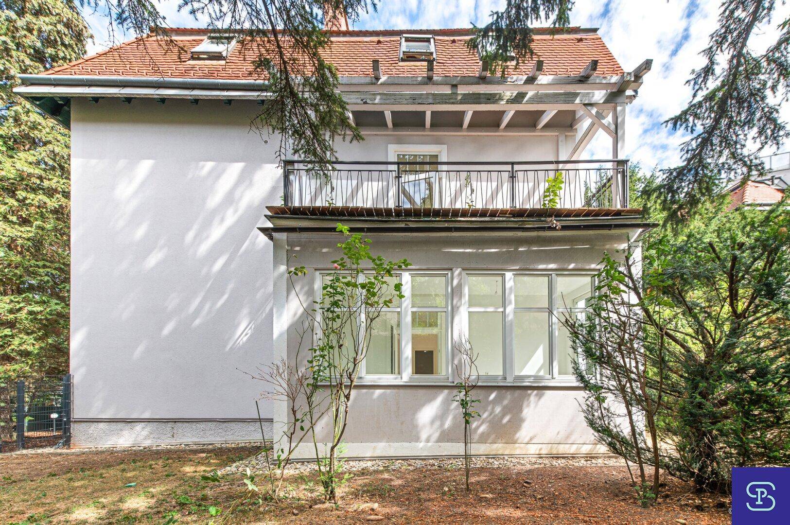 Rückansicht des Gebäudes mit Balkon und Veranda, umgeben von altem Baumbestand.