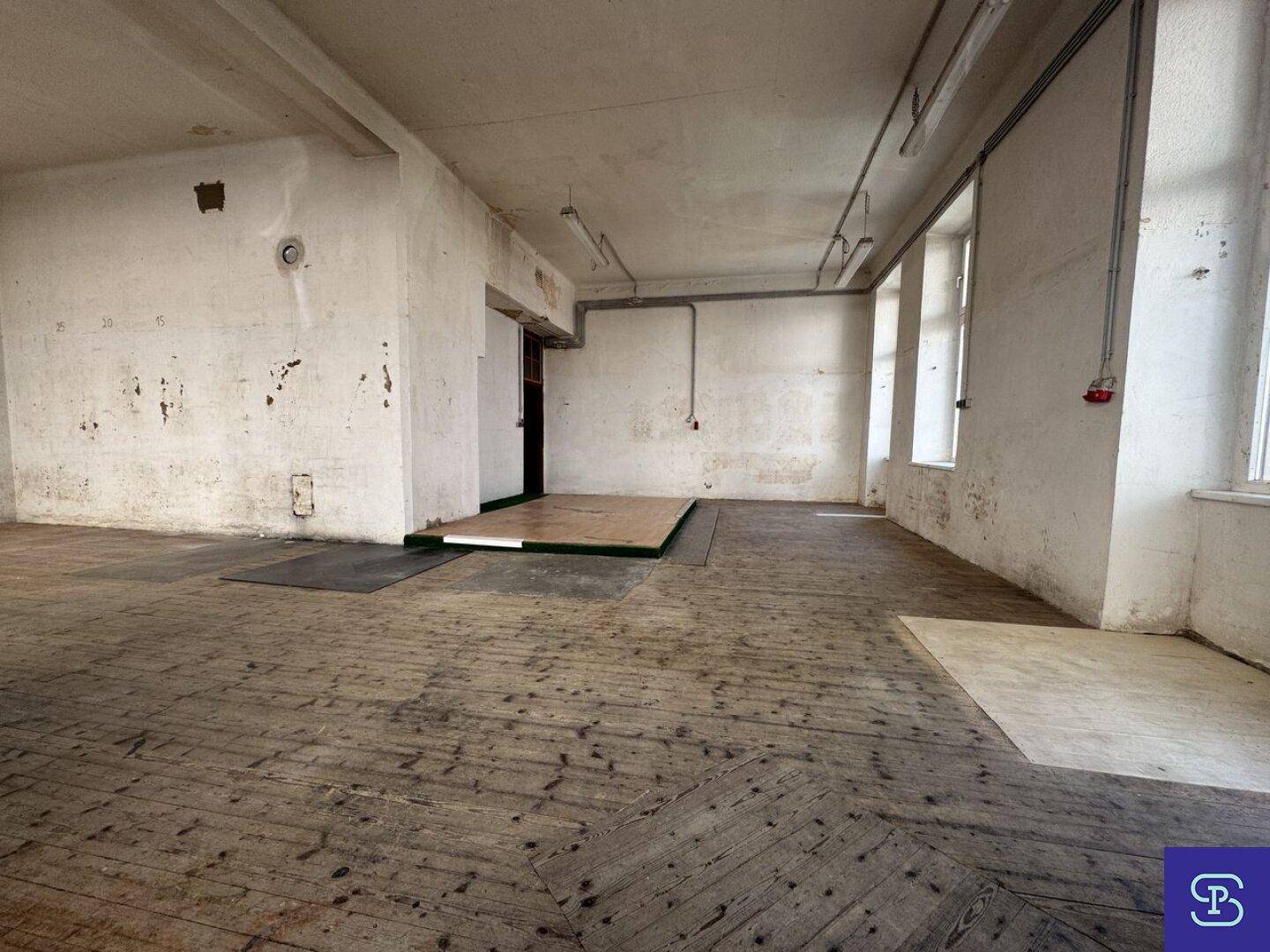Leerer Gewerberaum mit abgenutztem Holzboden und einer erhöhten Plattform in der Mitte des Raumes.