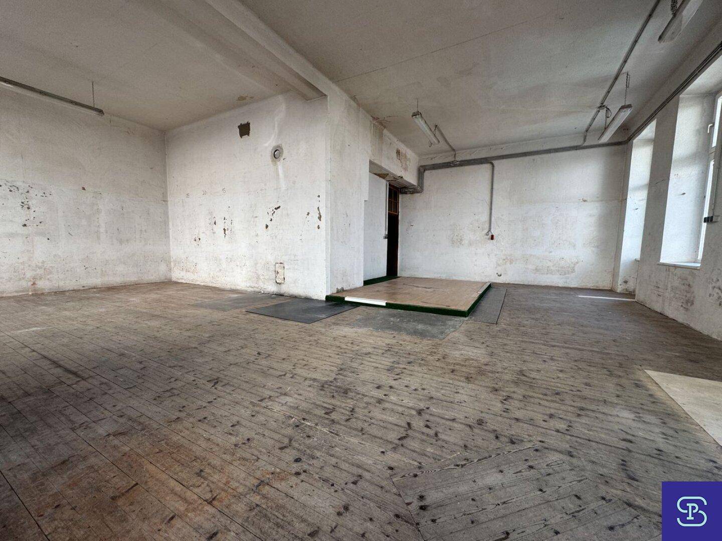 Geräumiger, unmöblierter Gewerberaum mit hohen Decken, großen Fenstern und sichtbaren Installationen.
