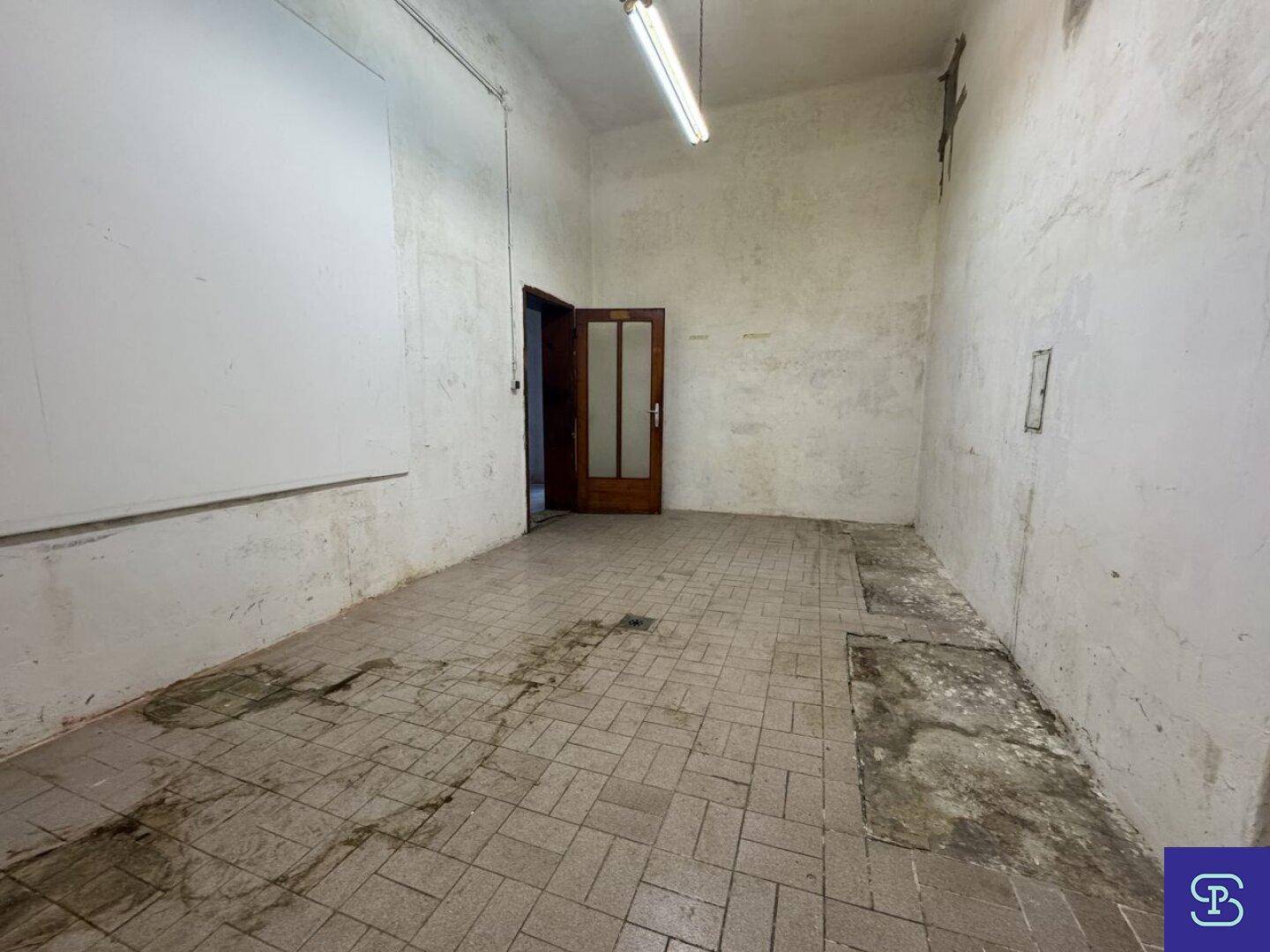 Leerer Raum mit gefliestem Boden, einer Holztür und einer großen weißen Wandfläche.