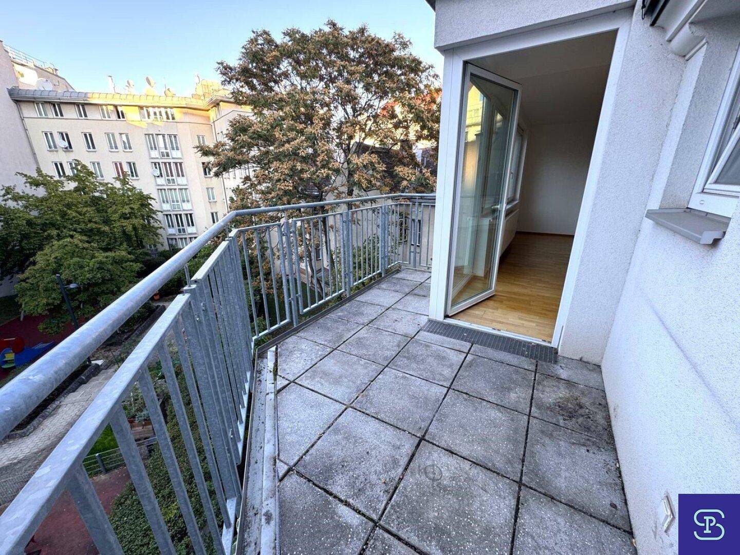 Balkon mit Fliesenboden und Zugang zum Innenraum, umgeben von Bäumen und Gebäuden.