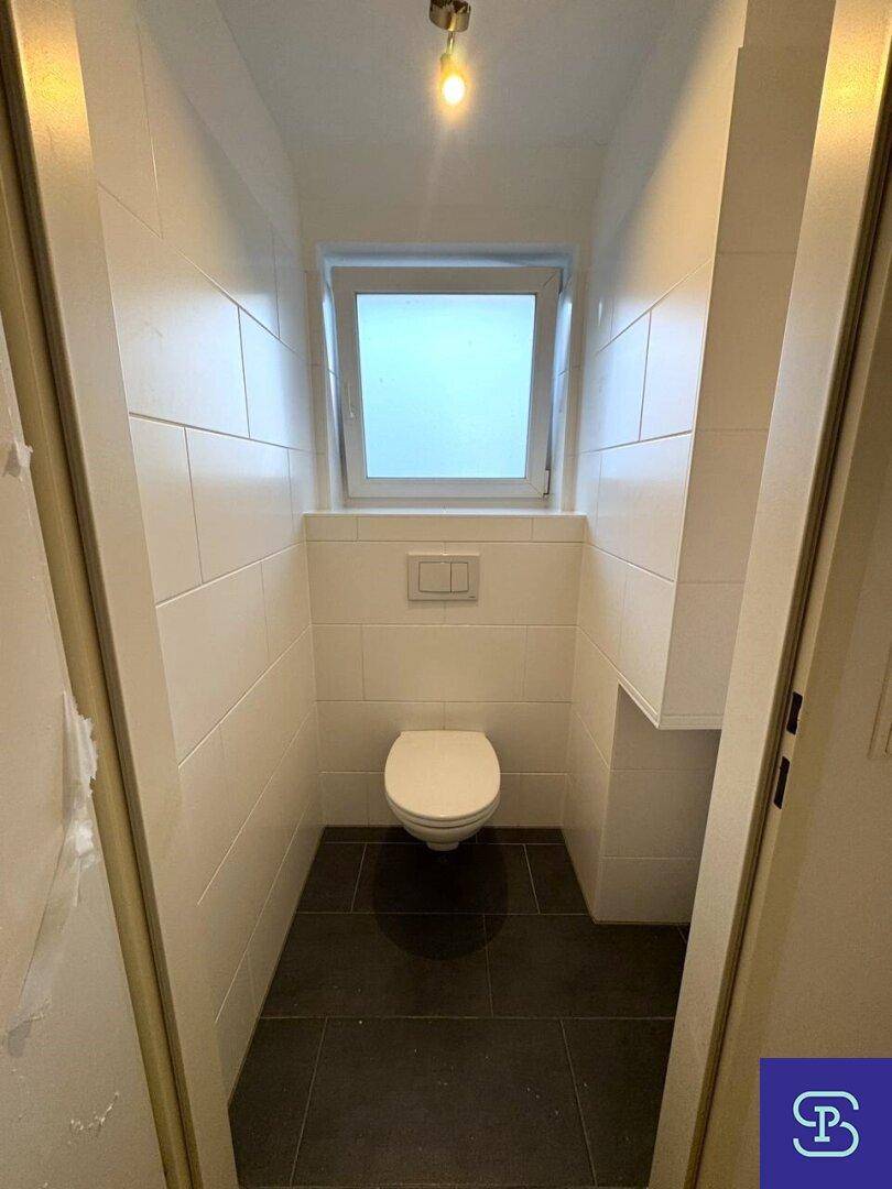 Separates WC mit wandhängender Toilette, weißen Fliesen und einem kleinen Fenster.