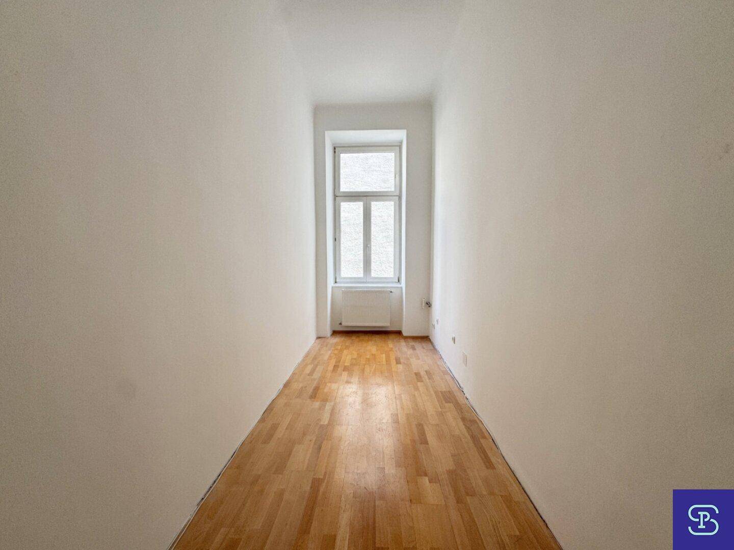 Langer, schmaler Raum mit Holzboden und einem Fenster am Ende, das Tageslicht spendet.