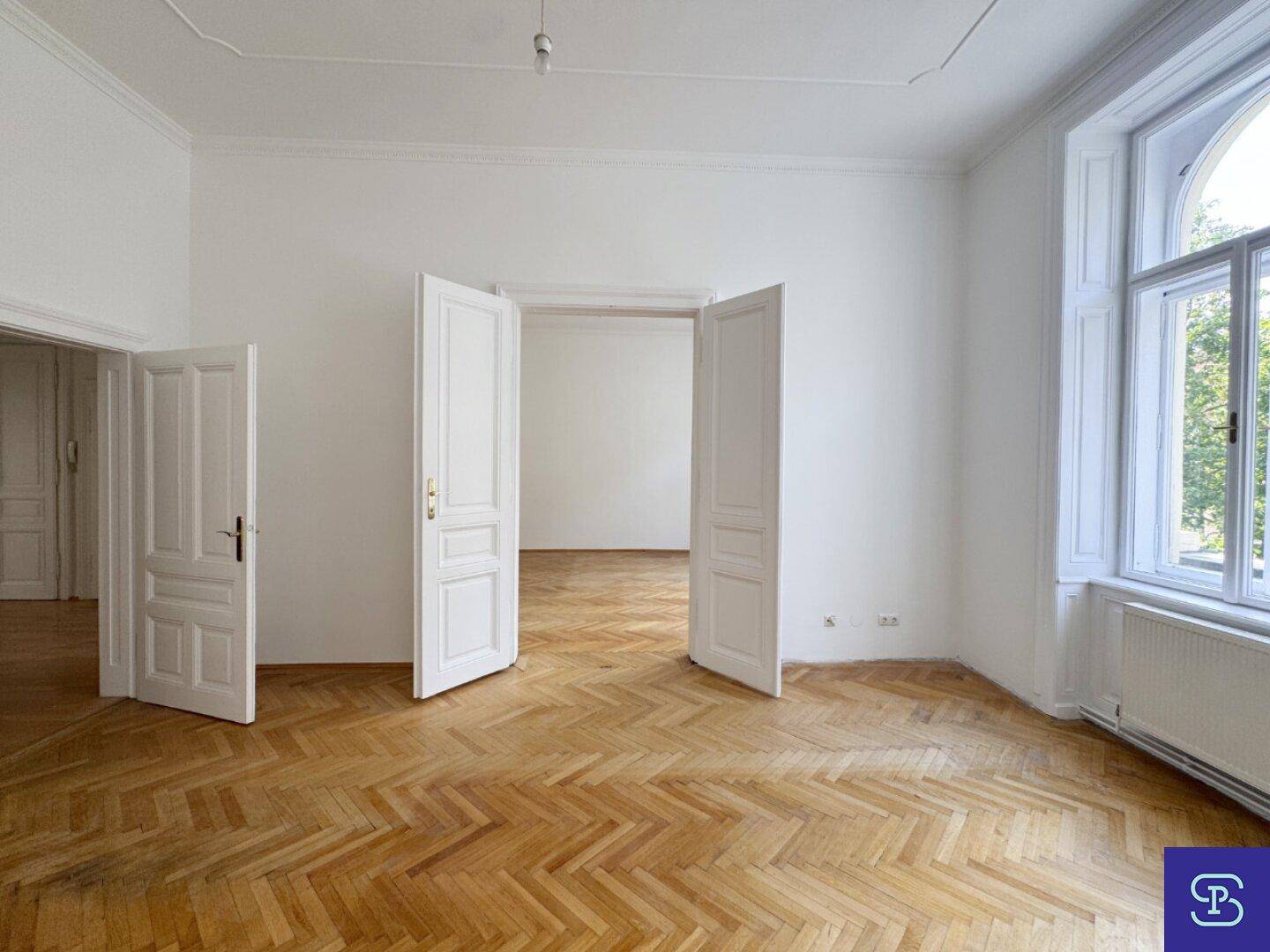 Helles Zimmer mit Fischgrätparkett, hohen Fenstern und weit geöffneten Flügeltüren.