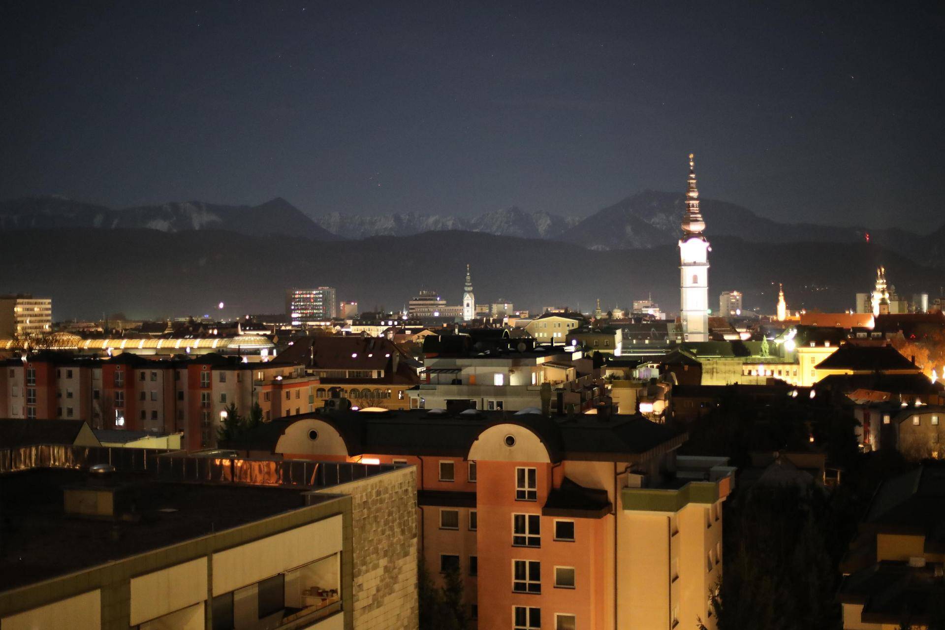 Nachtansicht der Stadt mit beleuchteten Gebäuden und schneebedeckten Bergen im dunklen Hintergrund.