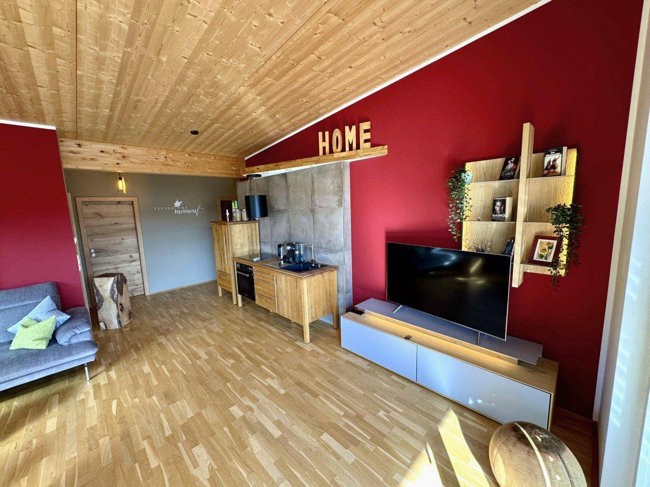 Wohnbereich mit kleiner Küchenzeile, roter Wand und Holzbalkendecke.