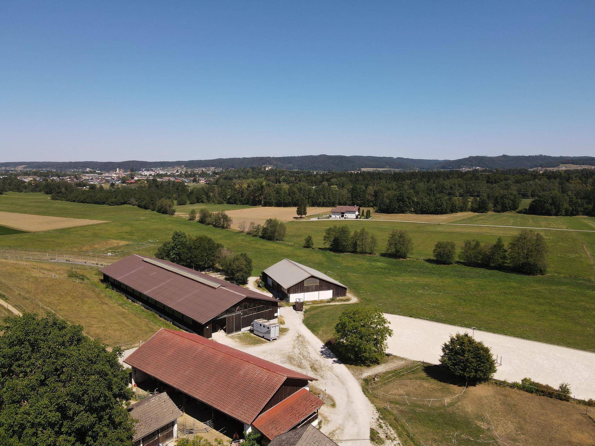Weitläufige Luftaufnahme eines Bauernhofs mit mehreren Gebäuden, Feldern und einer Reitbahn in ländlicher Umgebung.